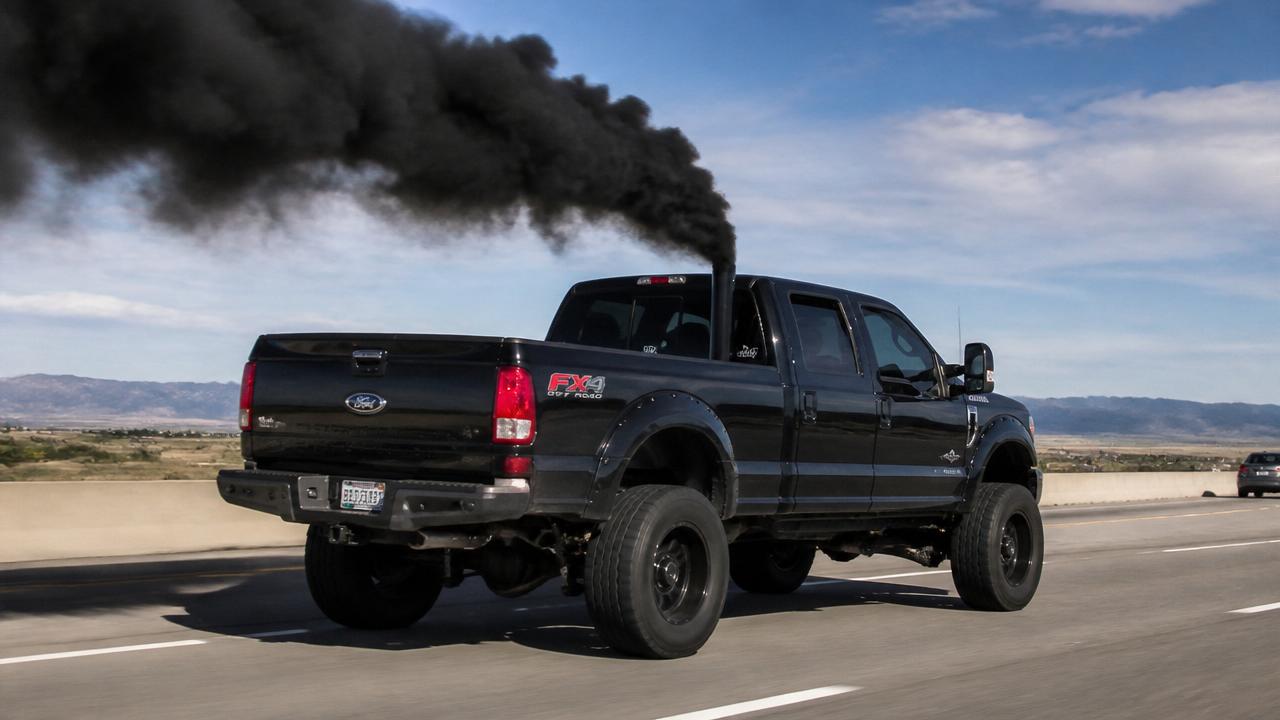 Modern black Ford F-250 dual cab pickup truck rolling coal with thick black soot smoke pouring from a vertical exhaust stack on a highway